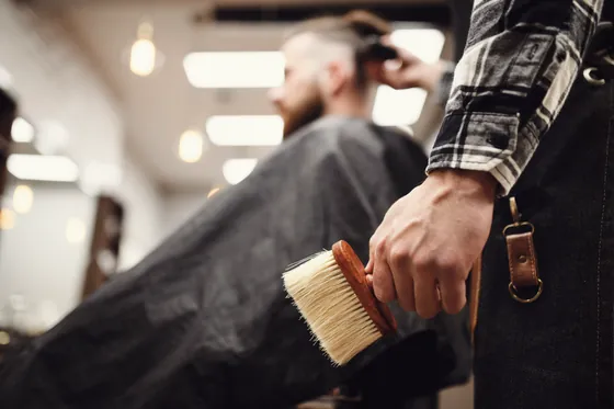 Stylist holding a neck brush beside a seated client in a cape.