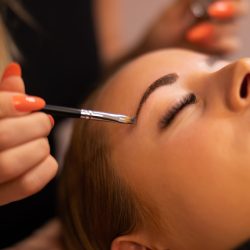 Hands of cosmetologist applying dye through brush while coloring female client's eyebrow at salon